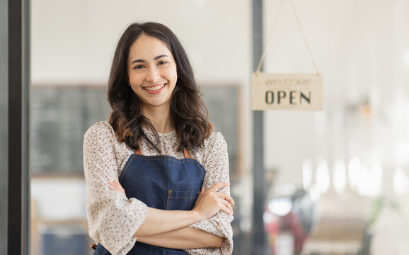 Portrait of happy waitress standing at restaurant entrance. Portrait of young business Asian woman attend new customers in her coffee shop. Smiling small business owner showing open sign in her shop.