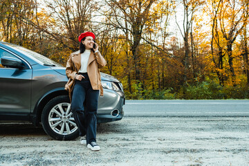 smiling woman in red beret talking on mobile phone leaning on parked car near autumn forest