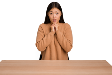 Young asian woman sitting on a table isolated praying for luck, amazed and opening mouth looking to front.