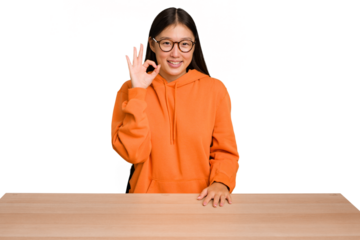 Young asian woman sitting on a table isolated winks an eye and holds an okay gesture with hand.