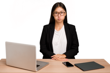 Young business asian woman sitting on a table isolated blows cheeks, has tired expression. Facial expression concept.