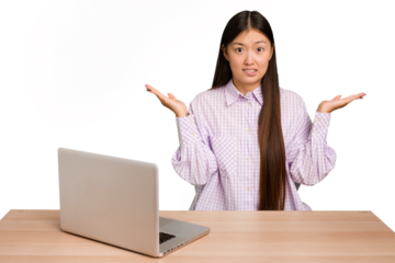 Young student asian woman in a workplace with a laptop isolated confused and doubtful shrugging shoulders to hold a copy space.