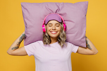Calm young smiling happy woman she wears pyjamas jam eye mask headphones listen tranquil music rest relax at home hold in hand sleep on pillow isolated on plain yellow background. Night nap concept.