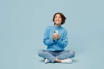 Full body fun young woman wear knitted sweater hold in hand use mobile cell phone look aside on workspace isolated on plain pastel light blue cyan background studio portrait. People lifestyle concept. - obrazy, fototapety, plakaty