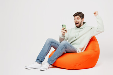 Full body young happy fun man wear mint hoody sit in bag chair hold in hand use mobile cell phone...