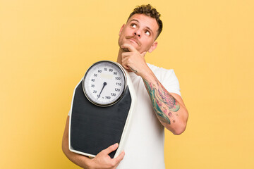 Young caucasian man holding a scale isolated looking sideways with doubtful and skeptical...