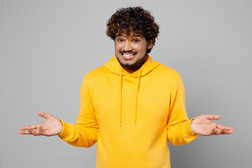 Young Indian man he wear casual yellow hoody shrugging shoulders looking puzzled, have no idea, nothing to say spread hands isolated on plain grey background studio portrait People lifestyle portrait