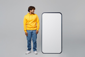 Full body smiling happy fun young Indian man wear casual yellow hoody stand near big huge blank screen mobile cell phone with workspace look at area isolated on plain grey background studio portrait.