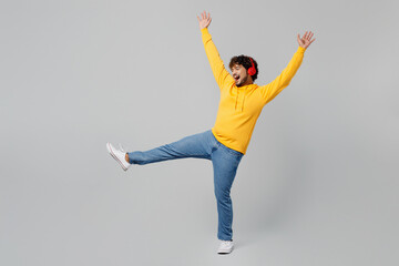 Full body young Indian man wear casual yellow hoody headphones listen to music dance raise up hands legs have fun on party isolated on plain grey background studio portrait. People lifestyle portrait.