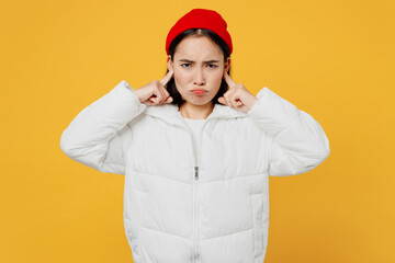 Young stressed sad woman of Asian ethnicity wear white padded windbreaker jacket red hat cover ears with hands fingers do not want to listen scream isolated on plain yellow background studio portrait.