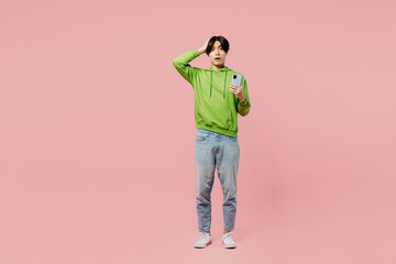 Full body shocked sad young man of Asian ethnicity wear green hoody look camera use mobile cell phone hold head isolated on plain pastel light pink background studio portrait People lifestyle concept