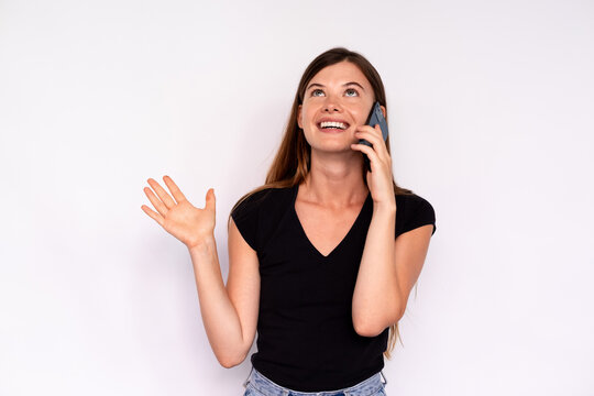 Portrait of excited young woman talking to friend on smartphone over white background. Caucasian lady wearing black T-shirt and jeans having mobile conversation. Mobile communication concept