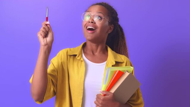 Young inspired cheerful African American woman raises hand with pen up having come up with idea for report at university and holds textbooks and workbooks stands in lilac studio. Education, training