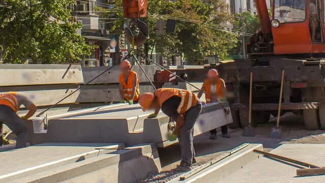 Installing concrete plates by crane at road construction site timelapse. Industrial workers with hardhats and uniform. Reconstruction of tram tracks in the city street. Blue clear sky
