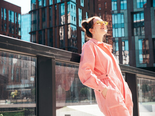 Young beautiful smiling hipster woman in trendy summer pink overalls. Carefree teen model posing in the street background at sunset. Positive female outdoors in sunglasses