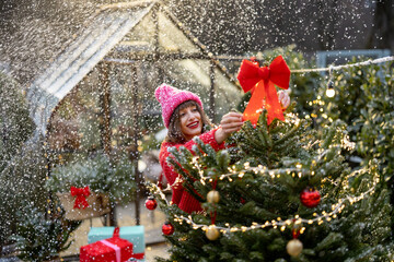 Young happy woman in red decorates lush Christmas tree with festive ballls and bows illuminated...