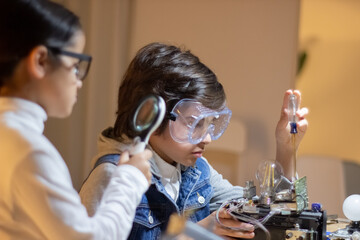 Kids geek creating new electronic device. Boy in protective glasses using screwdriver, girl looking...