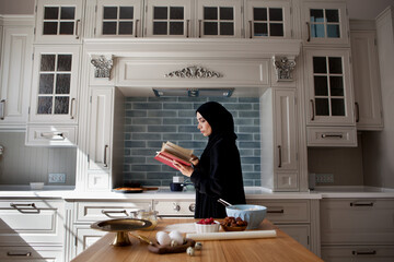 Beautiful Muslim woman cooks in kitchen and reads book, wears black abaya and hijab