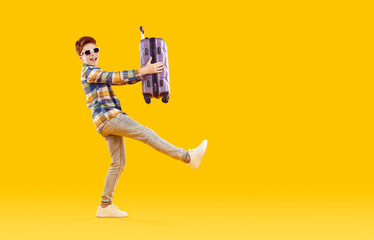 Fototapeta na wymiar Happy cute red-haired boy in sunglasses looks at the camera, dressed in bright plaid shirt and gray jeans, holding a suitcase in his hands. Studio full-size isolated shot on yellow background.