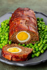 Sliced baked meatloaf made of ground meat, onion, carrot, stuffed with hard-boiled eggs, wrapped with bacon net, served with green peas. Festive food. Vertical image.