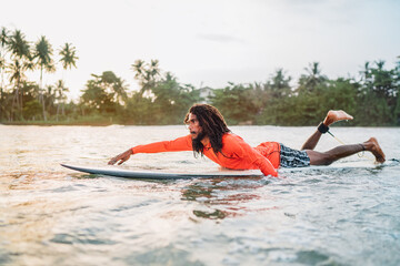 Black long-haired man paddling on long surfboard to the surfing spot in Indian ocean. Palm grove...