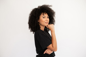 Thoughtful young woman holding thumb on her chin. Caucasian female model with afro hairstyle and brown eyes in black T-shirt standing sideways touching chin with thumb. Thought, dream concept - obrazy, fototapety, plakaty