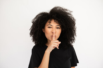 Fototapeta na wymiar Confident young woman making hush gesture. Caucasian female model with afro hairstyle and brown eyes in black T-shirt showing shh gesture telling something confidential. Secrecy, silence concept