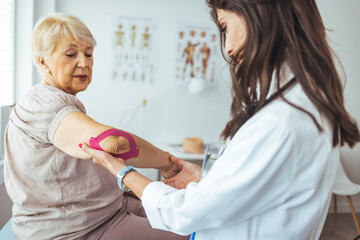 Fototapeta premium Kinesiology taping treatment with pink tape on senior patient injured arm. Woman hands apply kinesio treatment after muscle injury. A Modern rehabilitation physiotherapy worker with woman client