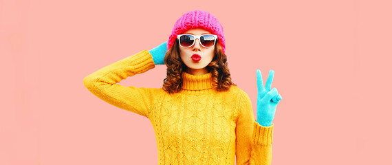 Winter portrait of cool woman blowing her red lips sending air kiss wearing colorful knitted yellow sweater, pink hat on background