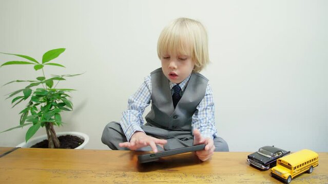 5 years old caucasian boy in grey suit and blue tie is playing computer game on tablet sitting at the desk in a room. Two toy cars on the table