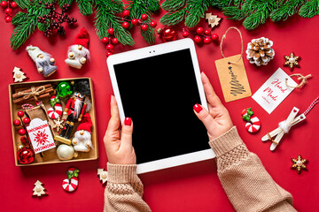 top view female hands holding tablet with black digital screen, Christmas box and decor on red table Flat lay Holiday shopping list, Happy New Year, online shop, chooses gifts, makes purchases Mockup - 553203761