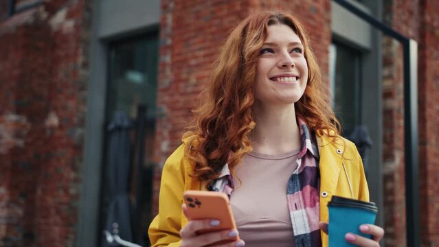 Happy young Caucasian woman with ginger hair holding blue paper cup of coffee and looking around. Joyful girl received message with good news on smartphone. Positive emotion. Outside. City life