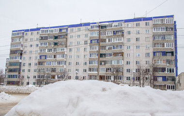 A large snowdrift in the background of a street with a multi-storey building. On the road lies white snow in high heaps. Urban winter landscape. Cloudy winter day, soft light.