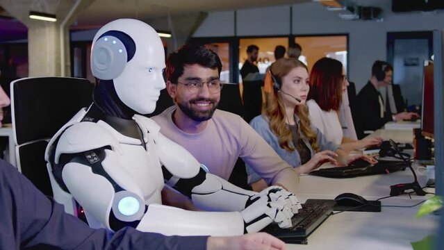 Side-view close-up portrait of white artificial intelligence robot working in office doing tasks with people. Futuristic cyborg typing on keyboard using computer. Workers watching bot working.