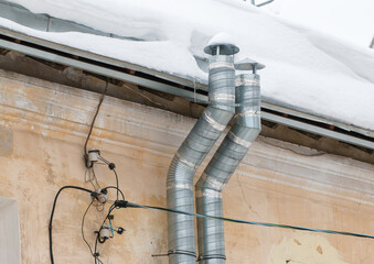 Two metal exhaust ventilation pipes on the wall of an old house. The roof of the building in white snow. Cloudy, cold winter day, soft light.