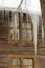 Large icicles hang on the edge of the roof, winter or spring. Log wall of an old wooden house with windows. Large cascades of icicles in smooth, beautiful rows. Cloudy winter day, soft light.