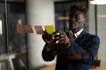 Portrait of successful businessman in office. Young African man witing ideas on colorful stickers on glass wall