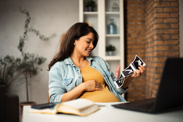 Beautiful young pregnant woman looking at ultrasound pictures of her baby. Businesswoman in office.