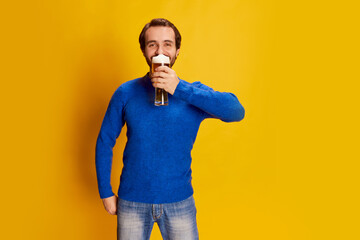 Portrait of emotive man in blue sweater posing with drink glass, cheerfully sipping beer isolated...