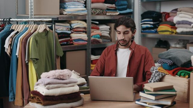 Attracrive disabled male manager working with laptop at the retail or boutique shop checking clothing stock in boxes on a checklist. Independent small business, store concept, entrepreneur