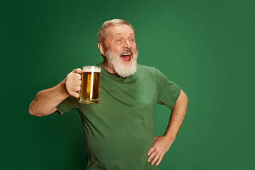 Portrait of senior man in T-shirt posing with beer isolated on green background. St Patrick's Day celebration. Drunk look - obrazy, fototapety, plakaty