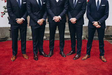 Wedding. Groom's friends. Business. A group of men in suits and leather shoes stand on a red carpet in front of the entrance to the building.