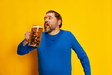 Portrait of emotive man in blue sweater posing with foamy beer mug, drinking with pleasure isolated...