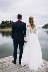 Wedding. The groom in a suit and the bride in a white dress are standing on the pier on the lake against the background of the sky and trees