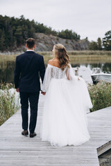 Wedding. The groom in a suit and the bride in a white dress are standing on the pier on the lake against the background of the sky and trees