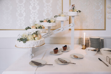 Wedding. Banquet. A cake for the bride and groom stands on a table with a white tablecloth, candles stand nearby and is decorated with flowers and greenery. Nearby are candles, plates and cutlery