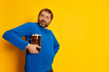 Portrait of emotive, funny man in blue sweater posing with foamy beer mug isolated over yellow...