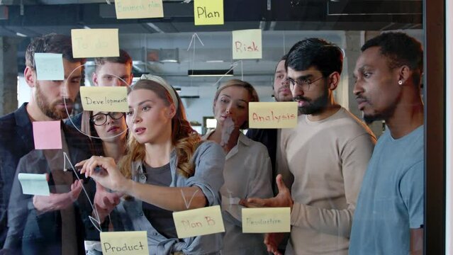 Portrait of focused professional IT workers brainstorming writing drawing on glass board coming up with ideas. Group of smart multiethnic colleagues standing in office with young woman in center.