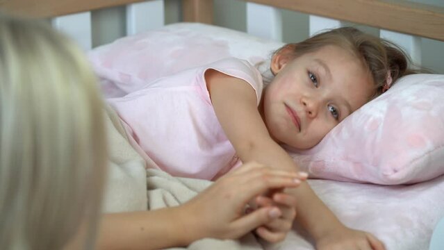 Mother sits next to little daughter in bed and puts sleep. Child laughs and naughty before bed. Healthy sleep concept