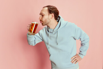 Portrait of young man in casual hoodie posing with lager foamy beer mug isolated over pink background. Favorite drink